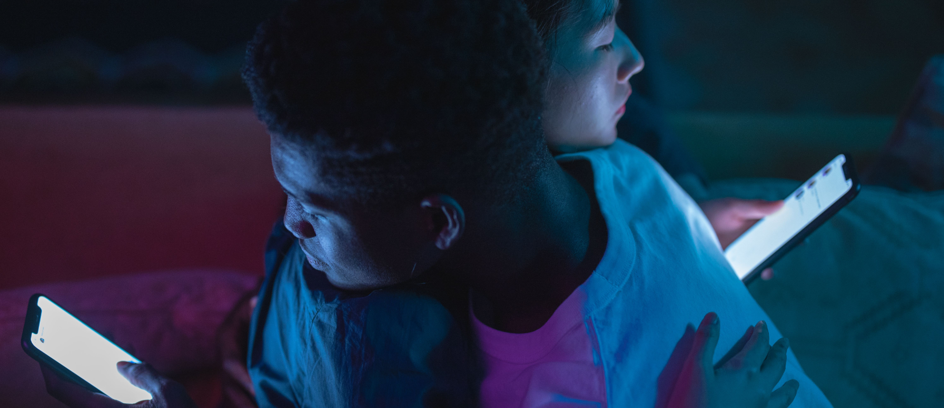 Dois corpos se abraçam na penumbra sob luzes neon azul e rosa, mas mantêm os olhos fixos nas telas brilhantes de seus respectivos smartphones por cima dos ombros.